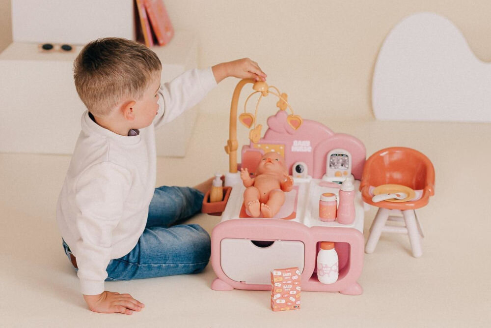 Chłopiec siedzący na podłodze bawi się różowym zestawem Baby Nurse z baldachimem. Lalka-niemowlę leży w łóżeczku z karuzelą.