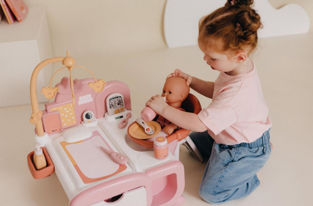 Dziewczynka bawi się zestawem Baby Nurse, karmiąc lalkę w różowym krzesełku do karmienia butelką. Widoczne akcesoria do pielęgnacji.