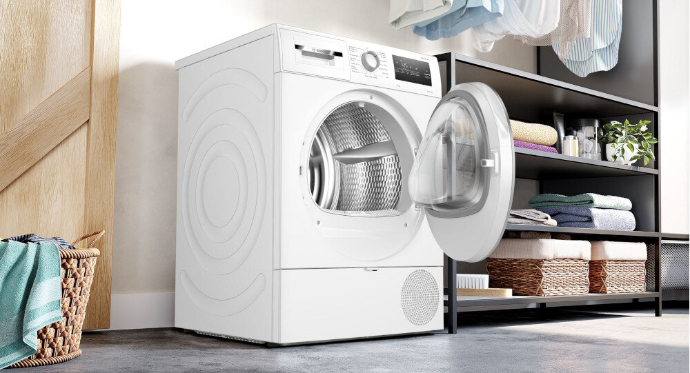 SUSZARKA BOSCH WTH85V9SPL Na zdjęciu widać suszarkę bębnową z otwartymi drzwiami. Urządzenie znajduje się w pralni lub garderobie. Po lewej stronie umieszczony jest kosz z ręcznikiem, a po prawej regał z poukładanymi tekstyliami, koszami oraz rośliną. Nad regałem wiszą ubrania. Wnętrze suszarki jest puste, przygotowane do załadunku. Całość wygląda czysto i nowocześnie cicha praca suszarki bosch redukcja hałasu antywibracje antivibration design stabilizacja bębna komfortowe suszenie minimalne drgania izolacja ścian amortyzator