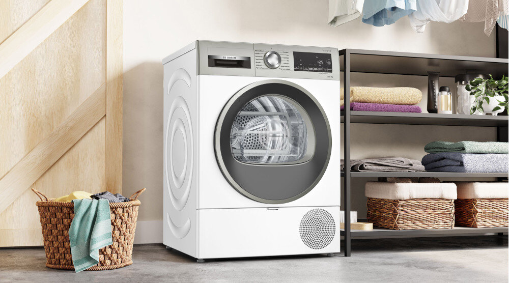 SUSZARKA BOSCH WQG243MSPL Na zdjęciu znajduje się suszarka do ubrań ustawiona we wnętrzu pralni lub garderoby. Obok niej stoi kosz wiklinowy z ręcznikami i ubraniami. Po prawej stronie widoczny jest regał z poukładanymi ręcznikami, tekstyliami oraz dekoracyjną rośliną doniczkową i koszami na dole półek. Na górze widać wiszące ubrania. Całość jest urządzone w jasnych, minimalistycznych kolorach, nadając pomieszczeniu czysty, schludny wygląd.
