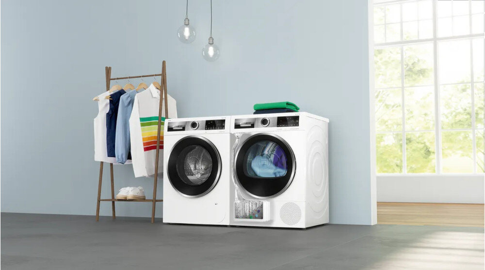 SUSZARKA BOSCH WQG243MSPL Na zdjęciu widać dwa urządzenia – pralkę i suszarkę ustawione obok siebie w jasnym, przestronnym pomieszczeniu. Po lewej stronie stoi stojak z powieszonymi ubraniami i parą butów, a na suszarce leżą złożone ręczniki. W tle duże okno z widokiem na zieleń, co dodaje wnętrzu jasności i świeżości. Całość prezentuje się czysto i minimalistycznie suszarka z pompą ciepła selfcleaning condenser automatyczne czyszczenie wymiennika ciepła energooszczędne suszenie wysoka efektywność suszenia dbałość o ubrania ekologiczne suszenie