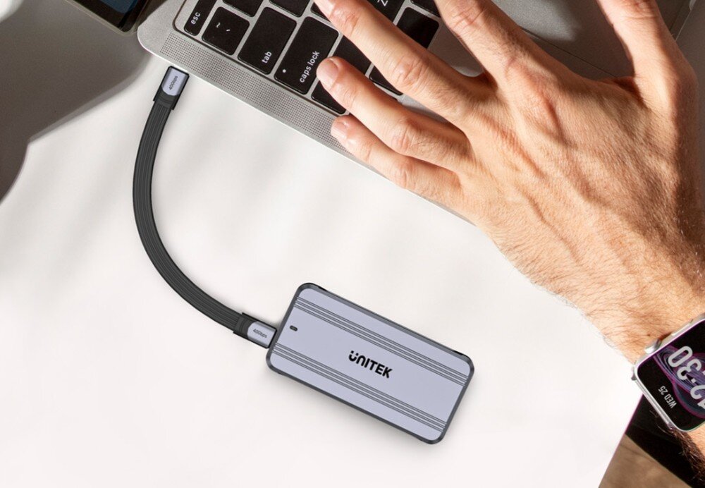 Obudowa dysku UNITEK S1242AGY01-EU Zbliżenie na dłoń osoby na klawiaturze srebrnego laptopa na jasnym blacie, z podłączonym kablem USB-C prowadzącym do srebrnej obudowy dysku, na której widnieje napis 'UNITEK', oraz z widocznym zegarkiem na nadgarstku