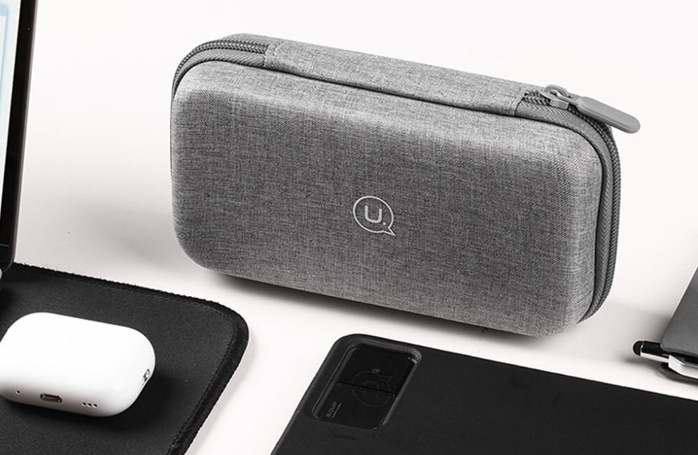 Szare etui ochronne z zamkiem błyskawicznym, z widocznym logo \'U\' na środku. Obok etui znajdują się inne akcesoria elektroniczne na białym tle.