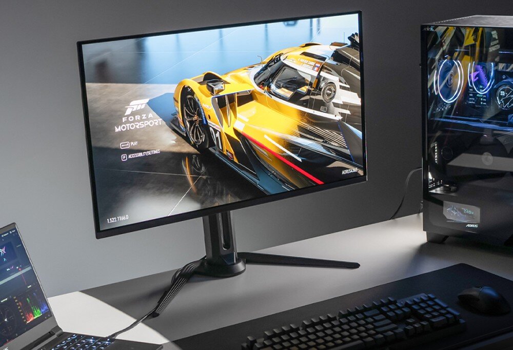 Monitor GIGABYTE Aorus FO27Q5P Monitor na biurku z włączoną grą wyścigową pokazuje żółty samochód, obok leży klawiatura, mysz oraz otwarty laptop i komputer stacjonarny
