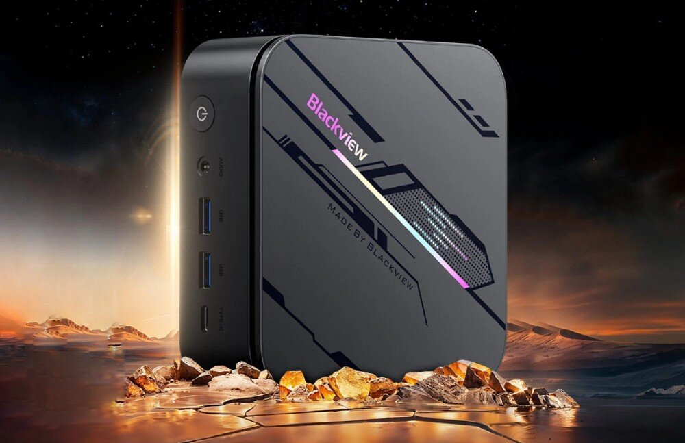 Komputer BLACKVIEW MP100 Pro Małe, czarne urządzenie o kwadratowym kształcie z logo 'Blackview' i kolorowym paskiem świetlnym, ustawione na kamieniach na tle zachodzącego słońca i krajobrazu pustynnego, opis