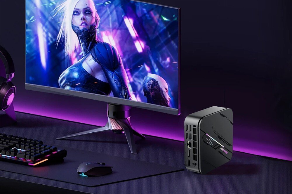 Komputer BLACKVIEW MP100 Pro Czarne miniaturowe urządzenie ustawione obok monitora z kolorową grafiką postaci w futurystycznym stylu, klawiatury i myszy na biurku podświetlonym na fioletowo, przeznaczenie, konstrukcja, wielkość