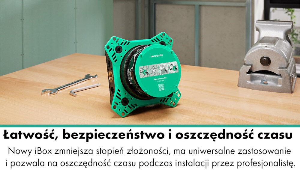 Na drewnianym stole znajduje się zielony element z napisem \'hansgrohe\' oraz \'Zestaw uniwersalny iBox\'. Obok leżą dwa klucze, a w tle widoczny jest imadło i ściana z metalowymi panelami.