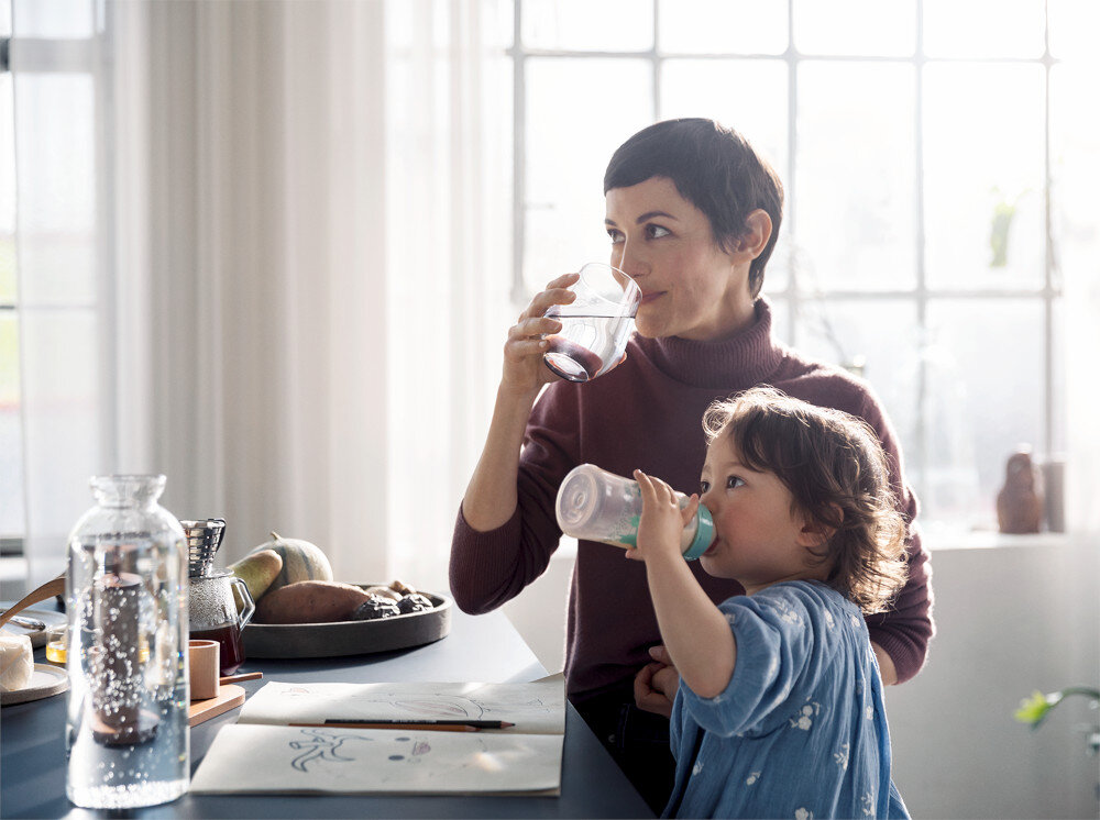 BATERIA KUCH HANSGROHE AQITTURA M91 Z FILTREM SODASYSTEM 210 76836000 Zdjęcie pokazuje kobietę stojącą przy kuchennym blacie, która pije napój z przeźroczystej szklanki, trzymając na rękach dziecko z butelką. Na blacie leżą kartki z rysunkami, kredki oraz szklana karafka i taca z warzywamiAqittura M91 SodaSystem 210 bateria kuchenna z sodą woda gazowana z kranu filtrowana woda mineralna ekologia bez plastiku oszczędność miejsca bateria kuchenna komfort w kuchni