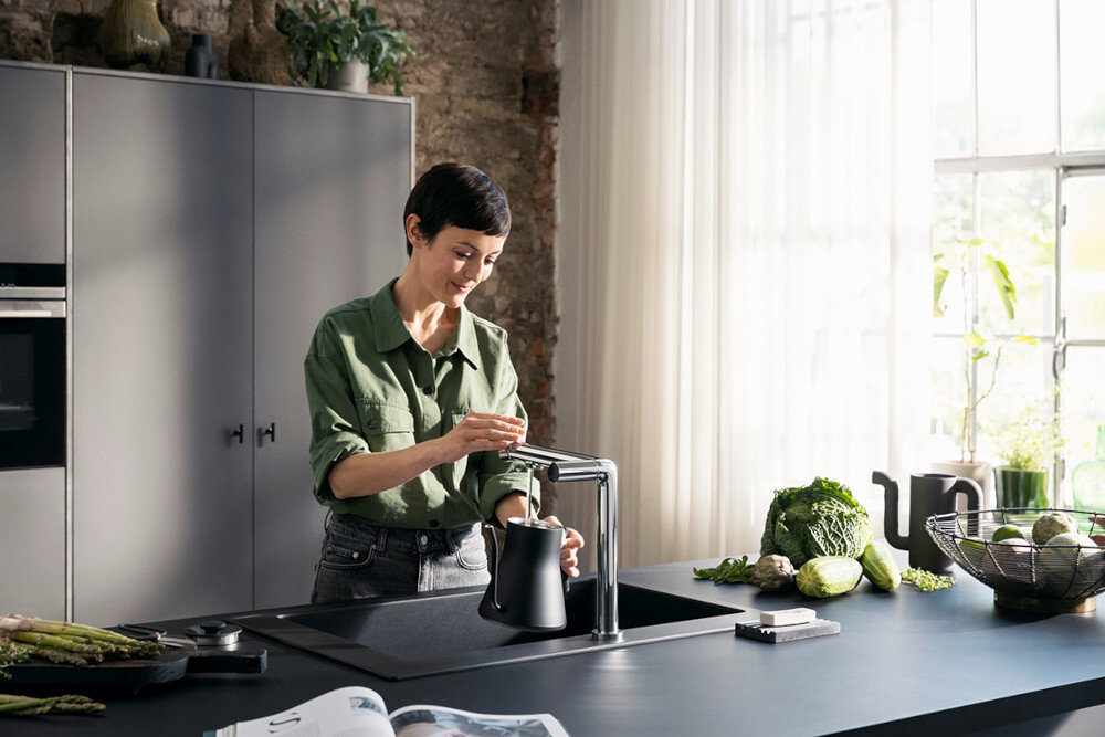 BATERIA KUCH HANSGROHE AQITTURA M91 Z FILTREM SODASYSTEM 210 76837000 Zdjęcie przedstawia jasną kuchnię z wyspą, w której centralnym punkcie jest chromowana bateria kuchenna przy prostokątnym zlewie. Na blacie leżą otwarta książka i tekstylia, a w tle widać zioła w doniczkach i misę z warzywami przy dużym oknie