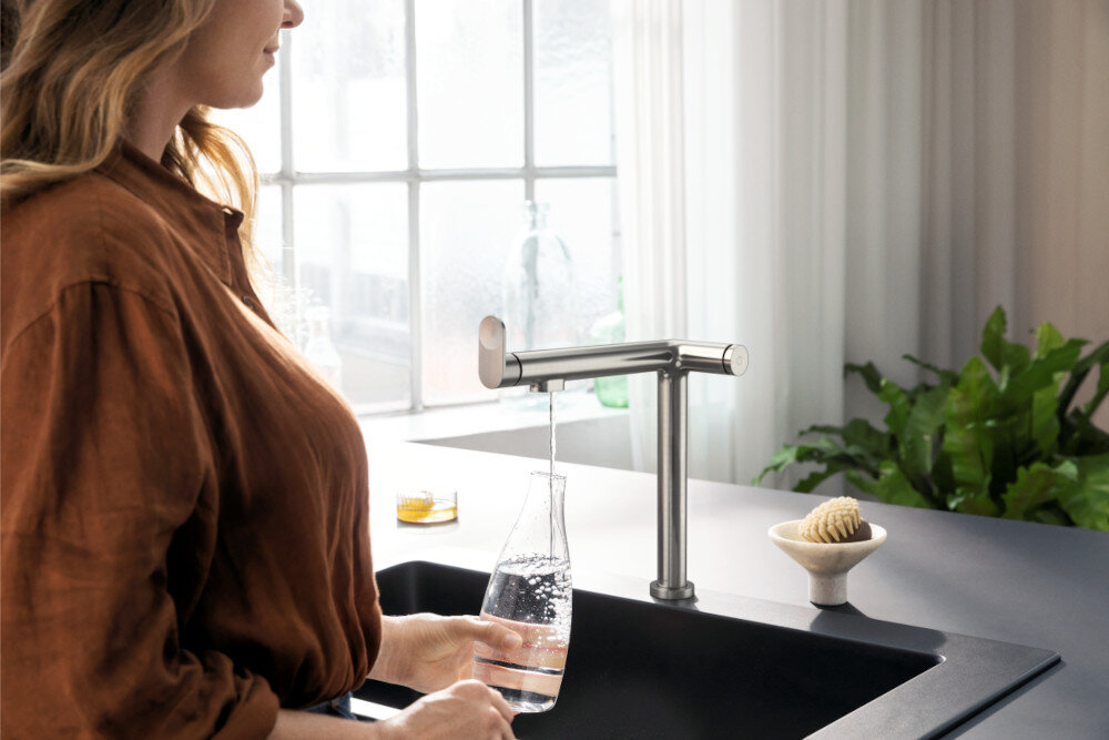 BATERIA KUCH HANSGROHE AQITTURA M91 Z FILTREM SODASYSTEM 210 76837800 Zdjęcie przedstawia jasną kuchnię z wyspą, w której centralnym punkcie jest Stal szlachetnaowana bateria kuchenna przy prostokątnym zlewie. Na blacie leżą otwarta książka i tekstylia, a w tle widać zioła w doniczkach i misę z warzywami przy dużym oknie