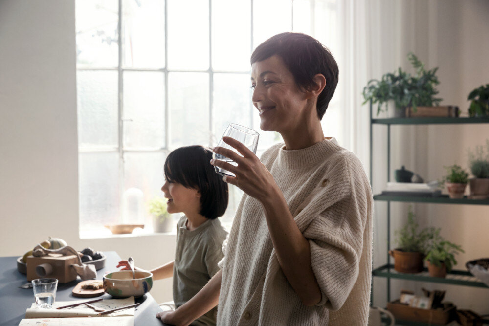 BATERIA KUCH HANSGROHE AQITTURA M91 Z FILTREM SODASYSTEM 210 76837800 Zdjęcie pokazuje kobietę stojącą przy kuchennym blacie, która pije napój z przeźroczystej szklanki, trzymając na rękach dziecko z butelką. Na blacie leżą kartki z rysunkami, kredki oraz szklana karafka i taca z warzywamiAqittura M91 SodaSystem 210 bateria kuchenna z sodą woda gazowana z kranu filtrowana woda mineralna ekologia bez plastiku oszczędność miejsca bateria kuchenna komfort w kuchni