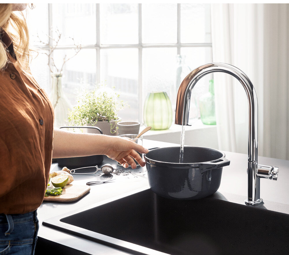 BATERIA KUCH HANSGROHE AQITTURA M91 Z FILTREM 210 76801000 Zdjęcie przedstawia jasną kuchnię z wyspą, w której centralnym punkcie jest chromowana bateria kuchenna przy prostokątnym zlewie. Na blacie leżą otwarta książka i tekstylia, a w tle widać zioła w doniczkach i misę z warzywami przy dużym oknie