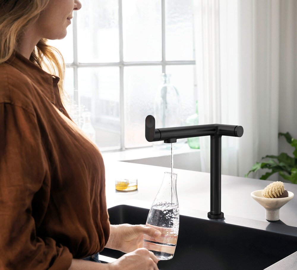 BATERIA KUCH HANSGROHE AQITTURA M91 Z FILTREM 210 76802670 Zdjęcie przedstawia jasną kuchnię z wyspą, w której centralnym punkcie jest Czarny matowana bateria kuchenna przy prostokątnym zlewie. Na blacie leżą otwarta książka i tekstylia, a w tle widać zioła w doniczkach i misę z warzywami przy dużym oknie