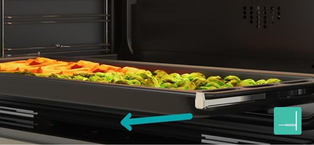 KUCHNIA GORENJE MEKS6141X Zbliżenie na blachę piekarnika wysuwaną na prowadnicach teleskopowych, na której pieką się pokrojone warzywa, a turkusowa strzałka u dołu wskazuje kierunek łatwego wysuwania