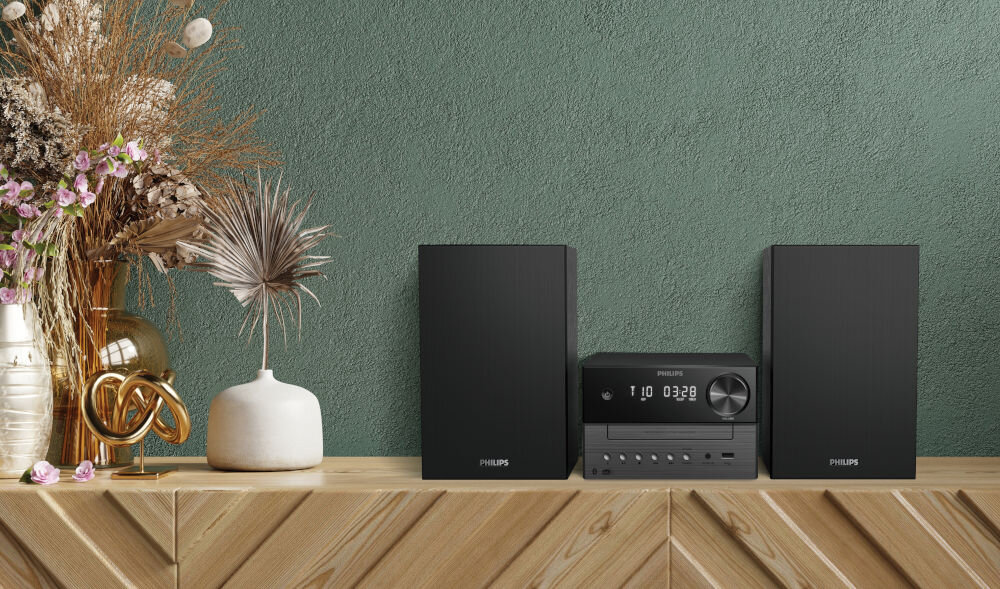 Na drewnianej półce stoi czarna wieża audio z dwoma głośnikami po bokach. Na wyświetlaczu wieży widoczny jest napis \'PHILIPS\' oraz cyfry \'03:29\'.