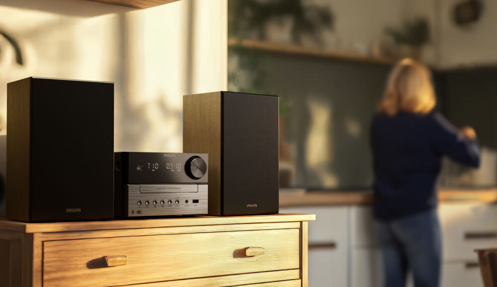 Na drewnianej komodzie stoi czarna wieża audio z dwoma głośnikami. W tle widoczna jest osoba w kuchni, ale nie jest ona w centrum uwagi.