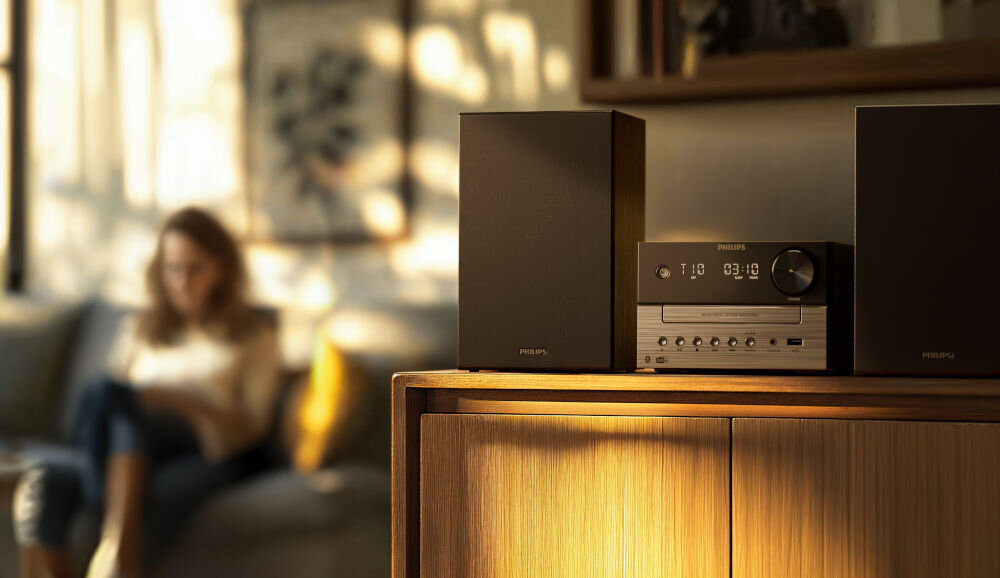 Na drewnianej szafce stoi czarna wieża audio z dwoma głośnikami, na której widoczne jest logo PHILIPS. W tle znajduje się osoba siedząca na sofie.