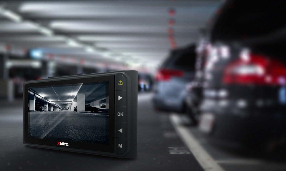 Wideorejestrator z logo \'xblitz\' wyświetla obraz z parkingu podziemnego. W tle widoczne są rozmyte sylwetki samochodów.