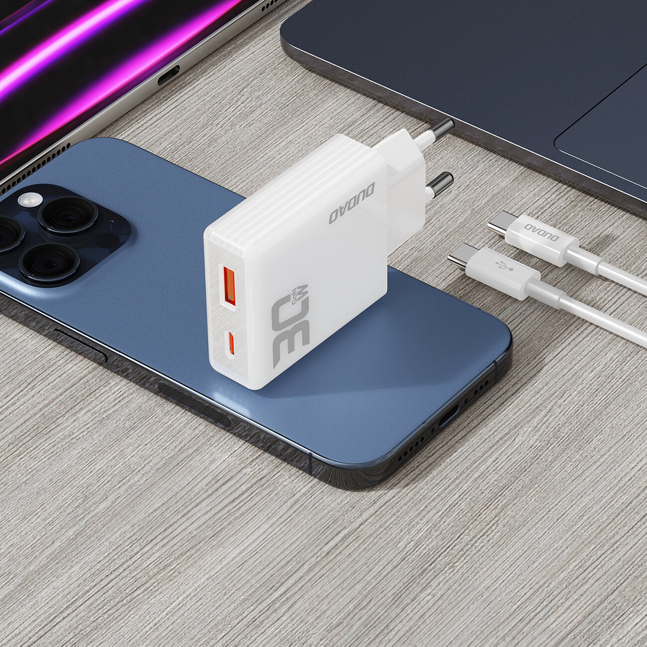 Na drewnianym blacie leży biały adapter z dwoma portami USB, oznaczony napisem \'Dudao\' i \'30W\'. Obok znajdują się dwa białe kable USB-C oraz smartfon i laptop.