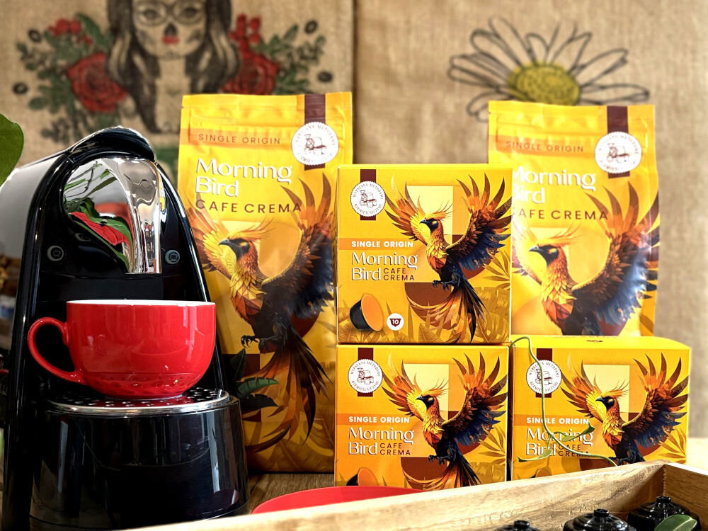 Kapsułki MORNING BIRD Cafe Crema 10 szt. i opakowania kawy ziarnistej w żółtych opakowaniaach na froncie ekspres kapsułkowy w tele ściana z brazyliskimi motywami