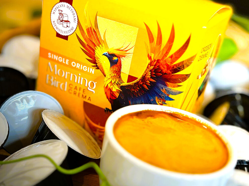 Kapsułki MORNING BIRD Cafe Crema 10 szt. w żółtym opakowaniu obok filiżanka z kawą i kilka kapsułek z kawą