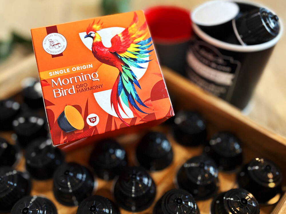 Kapsułki MORNING BIRD Oro Harmony 10 szt. w żółtym opakowaniu obok filiżanka z kawą i kilka kapsułek z kawą