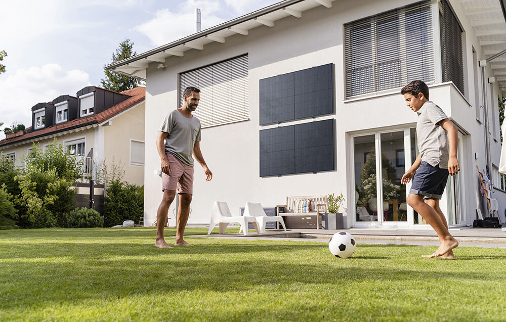 Fotowoltaika balkonowa ECOFLOW Mini Flex, zasilanie, bezpieczeństwo, bezpośrednio, wygenerować, w tle budynek i zamontowany do niego do ściany panele fotowoltaiczne, na pierwszym planie ojciec z synem grają w piłkę