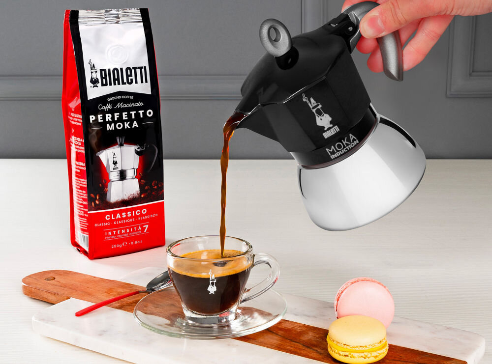 Osoba nalewa kawę z czarno-srebrnej kawiarki do szklanej filiżanki. Obok znajduje się opakowanie kawy z napisem \'Bialetti Perfetto Moka Classico Intensita 7\'.
