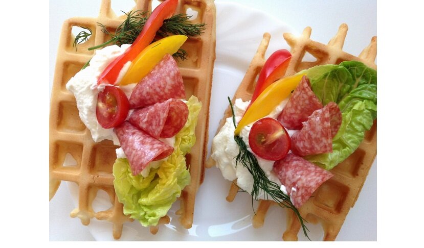 Dwa gofry na talerzu, udekorowane sałatą, plasterkami salami, pomidorkami koktajlowymi, papryką i koperkiem, z dodatkiem białego sosu.
