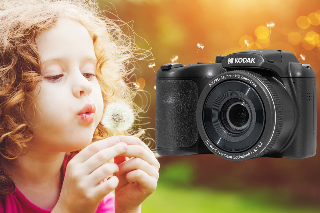 Na obrazie widoczny jest aparat fotograficzny z logo KODAK oraz dziecko dmuchające na dmuchawiec. Tło jest rozmyte, z ciepłymi kolorami i unoszącymi się nasionami dmuchawca.