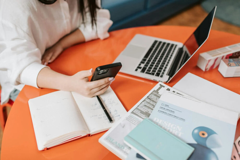 Urządzenie wielofunkcyjne BROTHER DCPT435WYJ1 Druk w kolorze, Wi-Fi kobieta siedząca przy stoliku z laptopem obok rozłożone notatki i dokumenty w dłoni trzyma telefon bezprzewodowe połączenie Wi-Fi wygoda pracy bez kabli swoboda w wyborze miejsca ustawienia