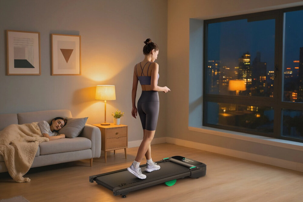 Bieżnia elektryczna UREVO Spacewalk 5 600W 6 km-h pokój osoba trenuje na bieżni obok na kanapie śpi druga osoba w tle okno z widokiem na miasto trening w domu zdrowie kondycja