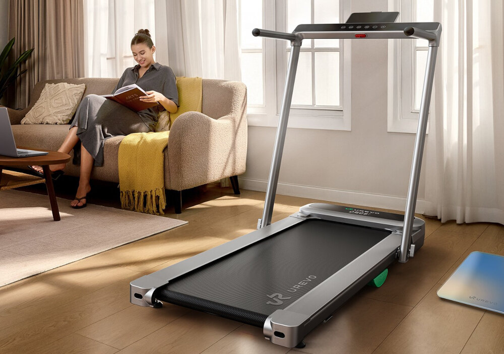 Bieżnia elektryczna UREVO Foldmix 5 Pro 1864W 13.4 km-h pokój bieżnia stoi na podłodze w tle na kanapie siedzi osoba czyta książkę okna meble trening domowy cardio kondycja zdrowie