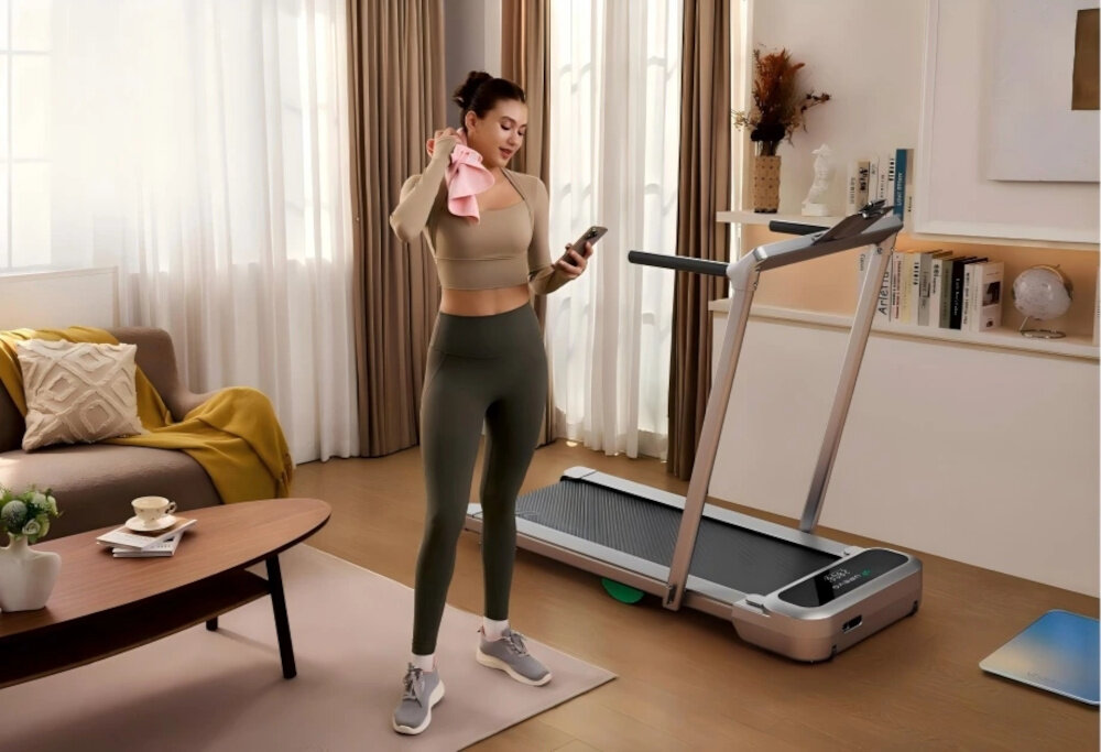 Bieżnia elektryczna UREVO Foldmix 5 Pro 1864W 13.4 km-h pokój osoba stoi z telefonem w dłoni wyciera się ręcznikiem obok stoi bieżnia w tle okna meble łączność Bluetooth aplikacja UREVO dostęp do realistycznych map tras trybów sportowych statystyk monitorowanie postępów wyznaczanie konkretnych celów treningowych na systemy Android i iOS