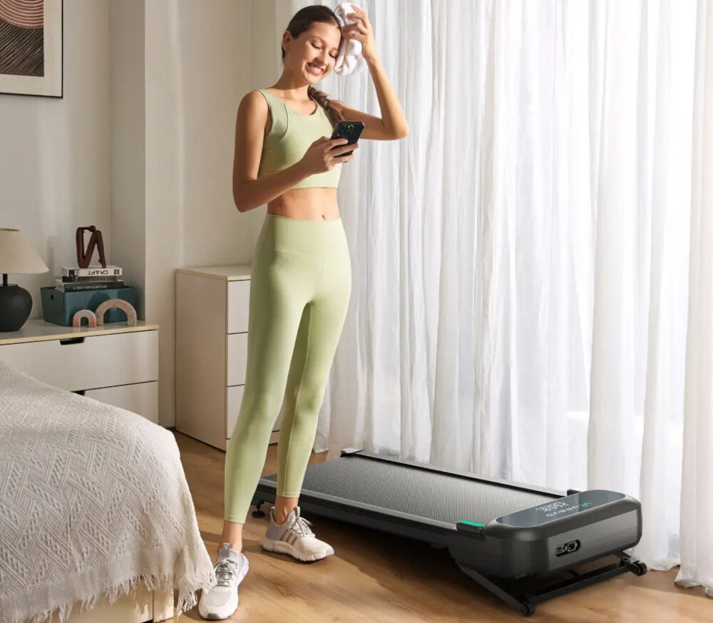 Bieżnia elektryczna UREVO Spacewalk 5L 600W 6 km-h pokój osoba stoi obok bieżni trzyma telefon w dłoni w tle okno trening w domu zdrowie kondycja