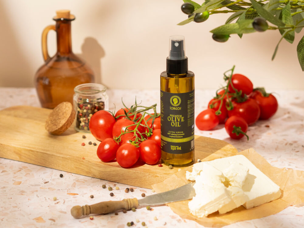 Oliwa z oliwek IORGOS Extra virgin z Kalamaty 150 ml Butelka oliwy z oliwek w sprayu marki Iorgos stoi na drewnianej desce do krojenia, otoczona pęczkiem pomidorków koktajlowych na gałązce, słoikiem z ziarnami pieprzu i kawałkami białego sera. W tle znajduje się bursztynowa karafka, a tło jest nakrapiane.