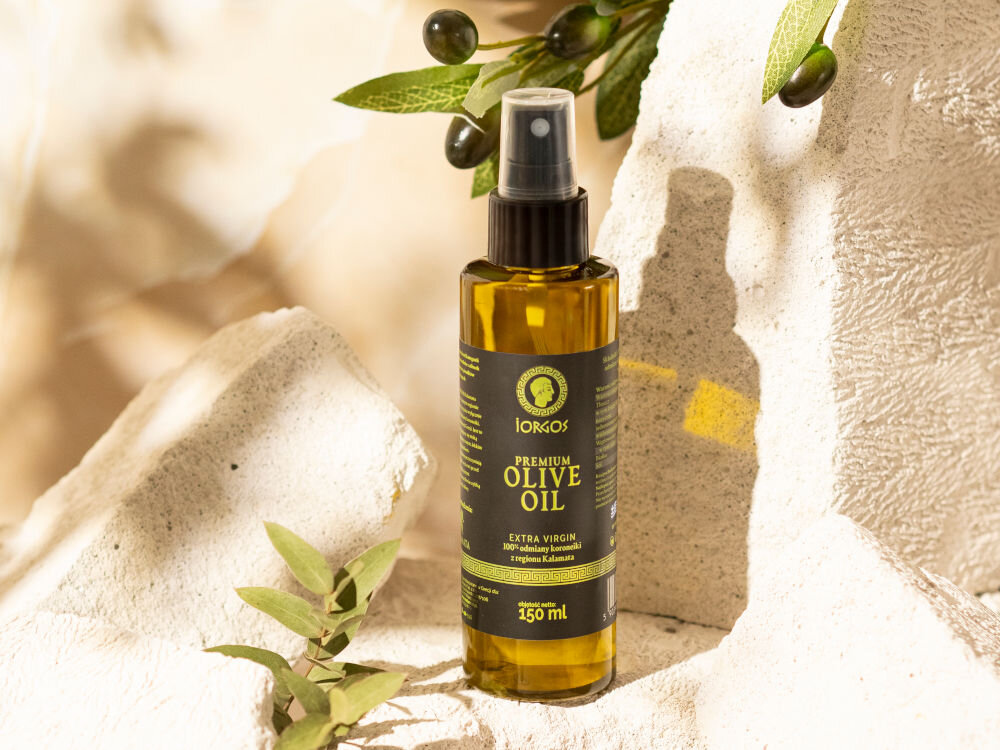 Oliwa z oliwek IORGOS Extra virgin z Kalamaty 150 ml Butelka oliwy z oliwek w sprayu marki Iorgos stoi na bloku z jasnego, porowatego kamienia. Obok butelki rośnie gałązka z liśćmi, a nad nią zwisa gałązka oliwna z ciemnymi oliwkami. Tło jest jasne i beżowe.