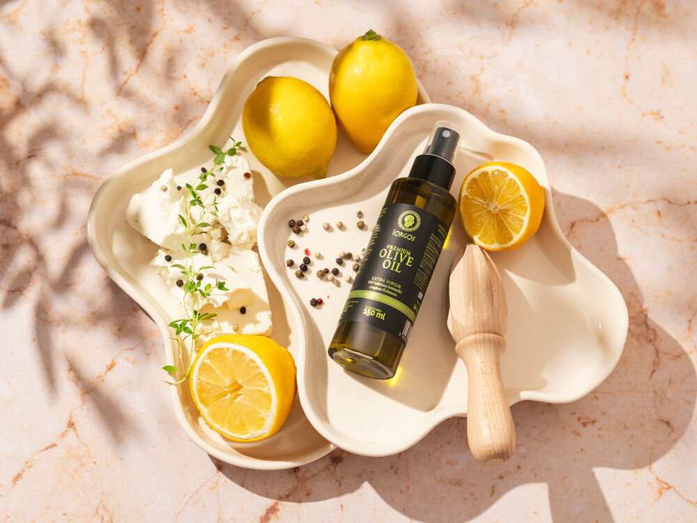 Oliwa z oliwek IORGOS Extra virgin z Kalamaty 150 ml Butelka oliwy z oliwek w sprayu marki Iorgos leży na kremowym, rzeźbionym talerzu obok dwóch całych cytryn, przekrojonej cytryny z wyciskaczem do cytrusów oraz kawałków białego sera z gałązkami tymianku i rozsypanymi ziarnami pieprzu. Tło jest marmurowe z cieniami liści.