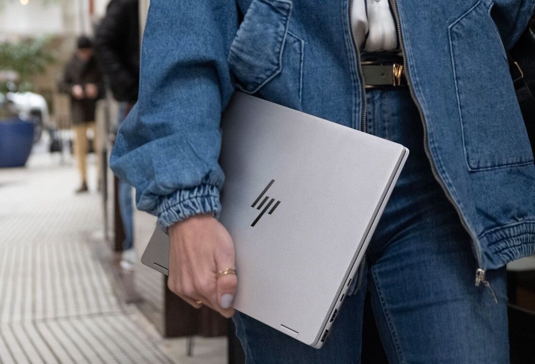 Osoba trzyma srebrny laptop z widocznym logo \'hp\'. Ubrana jest w niebieską kurtkę jeansową i dżinsy.