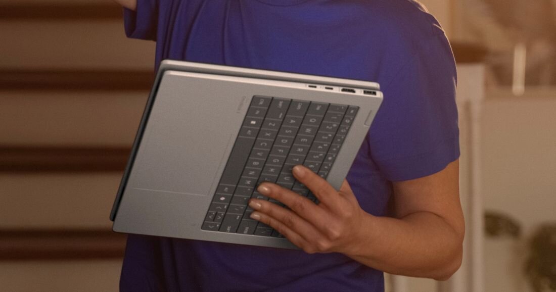Osoba trzyma laptopa w pozycji zamkniętej, widoczna jest klawiatura i porty boczne urządzenia. Laptop ma srebrną obudowę i ciemną klawiaturę.