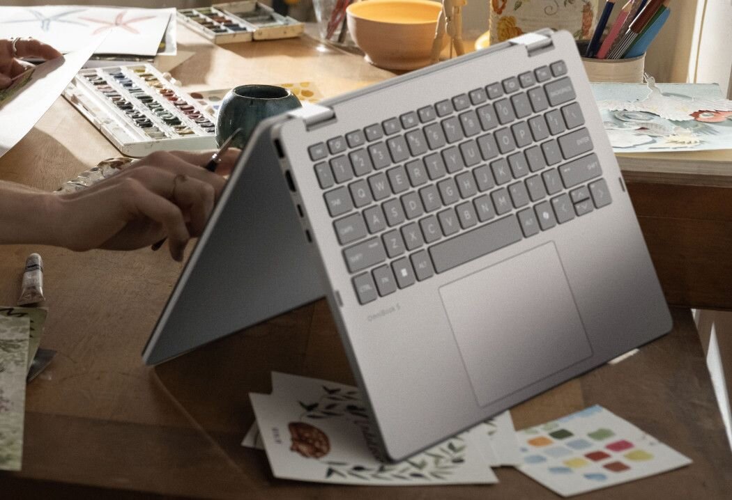 Laptop w trybie namiotu stoi na drewnianym stole, obok materiałów artystycznych i akcesoriów do malowania. Na klawiaturze widoczny jest napis \'OmniBook\'.