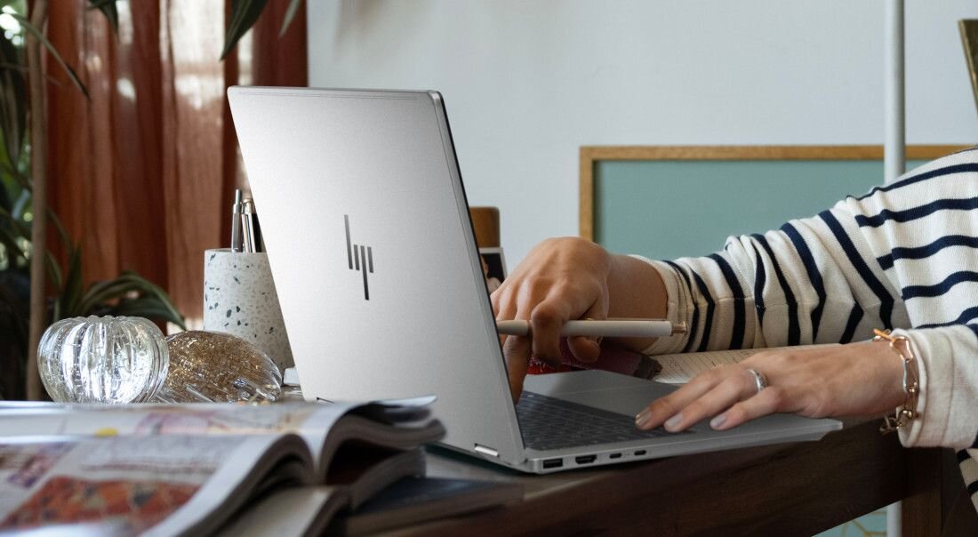 Osoba używa srebrnego laptopa z widocznym logo producenta na obudowie. Na stole obok znajdują się czasopismo i dekoracyjne przedmioty.