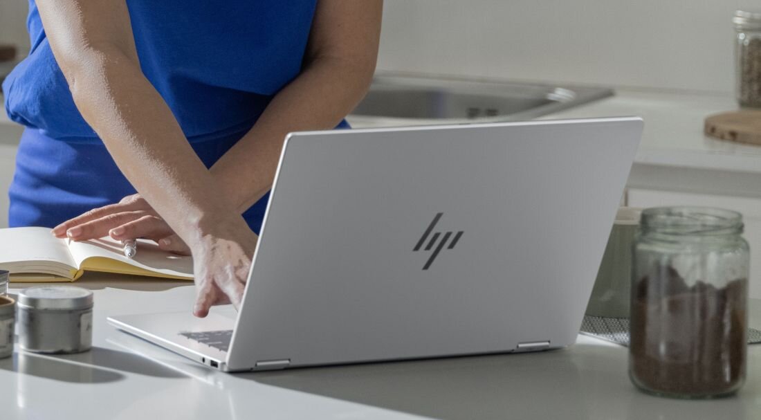 Osoba w niebieskim ubraniu stoi przy białym laptopie z logo HP, obok otwartej książki na blacie kuchennym.