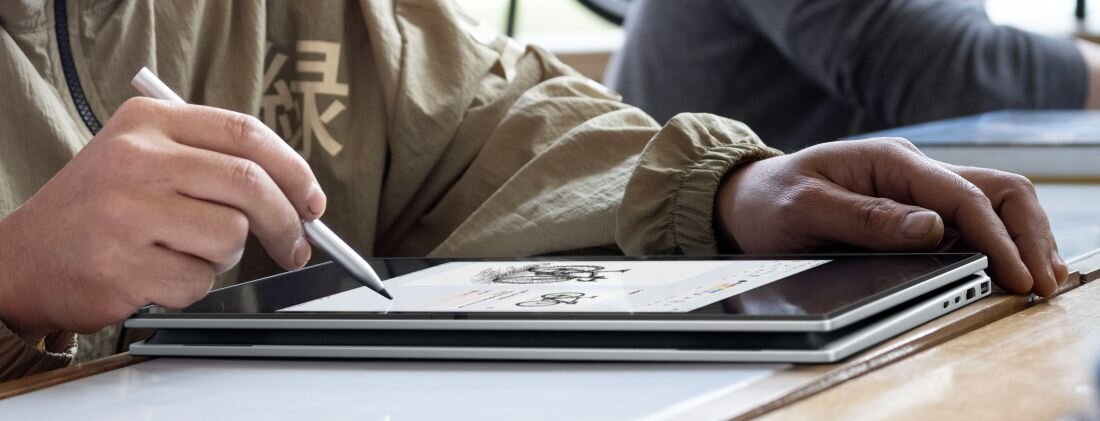 Osoba używa rysika na ekranie laptopa w trybie tabletu, na którym widoczny jest rysunek. Laptop leży na drewnianym stole, a osoba ma na sobie jasną kurtkę z widocznymi znakami.