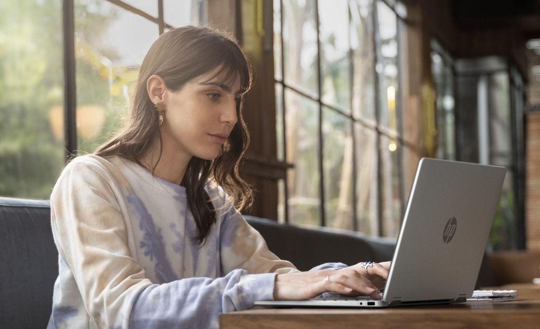 Osoba siedzi przy stole, używając laptopa z widocznym logo HP. W tle znajdują się duże okna z widokiem na zieleń.