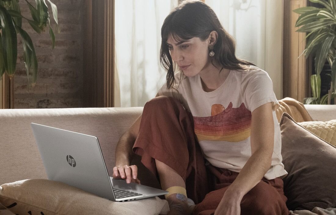 Osoba siedzi na kanapie, używając laptopa z widocznym logo HP. W tle znajdują się rośliny i zasłony.