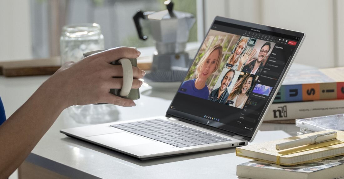 Osoba trzymająca kubek siedzi przy stole, na którym stoi otwarty laptop z widocznym ekranem pokazującym wideokonferencję. Obok laptopa leżą książki i notatnik z długopisem.