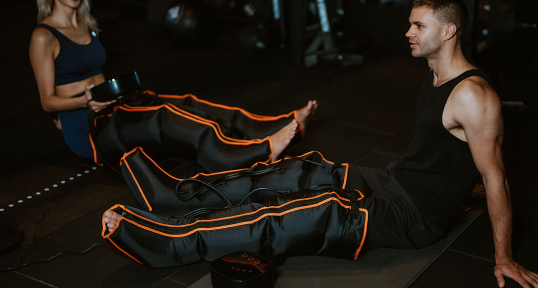 Dwie osoby siedzą na macie, mają na nogach urządzenia przypominające masażery limfatyczne z czarnym materiałem i pomarańczowymi paskami. W tle widoczne są elementy siłowni.