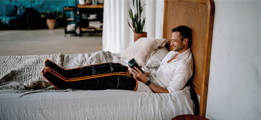 Osoba siedzi na łóżku z założonymi na nogi czarnymi masażerami z pomarańczowymi paskami, trzymając w ręku urządzenie sterujące. W tle widać roślinę i półkę z książkami.