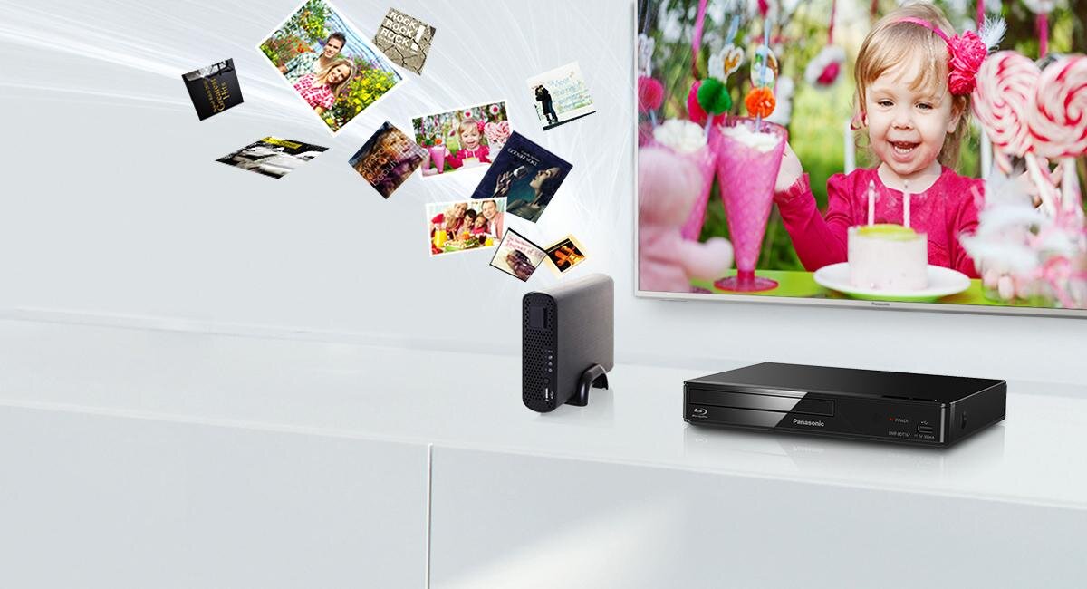 Na białej szafce znajduje się odtwarzacz Blu-ray z logo Panasonic oraz małe urządzenie elektroniczne. W tle widać telewizor z wyświetlonym zdjęciem dziecka przy torcie, a wokół unoszą się różne zdjęcia.